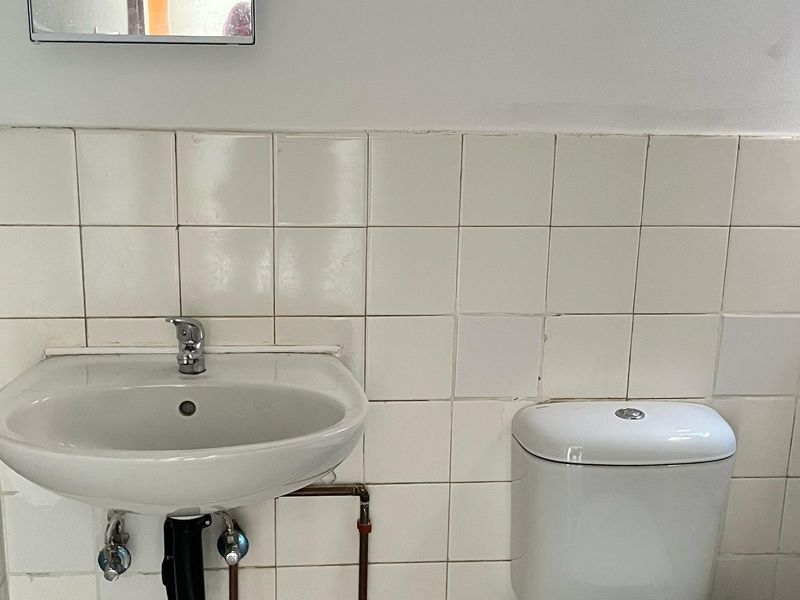 A functional bathroom featuring a pedestal washbasin, toilet, and white ceramic wall tiles with exposed plumbing.