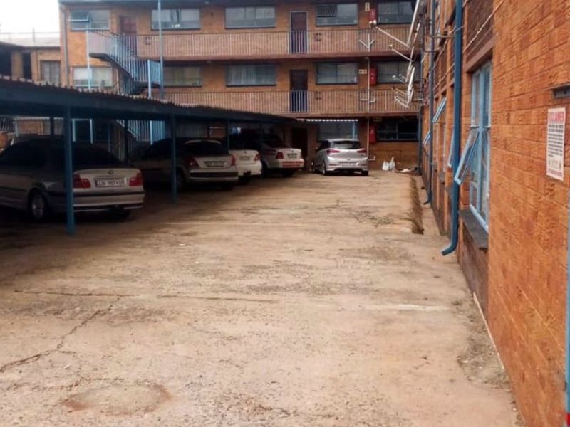 Exterior view of an apartment complex featuring a paved parking area with covered bays and multi-story brick building facade.