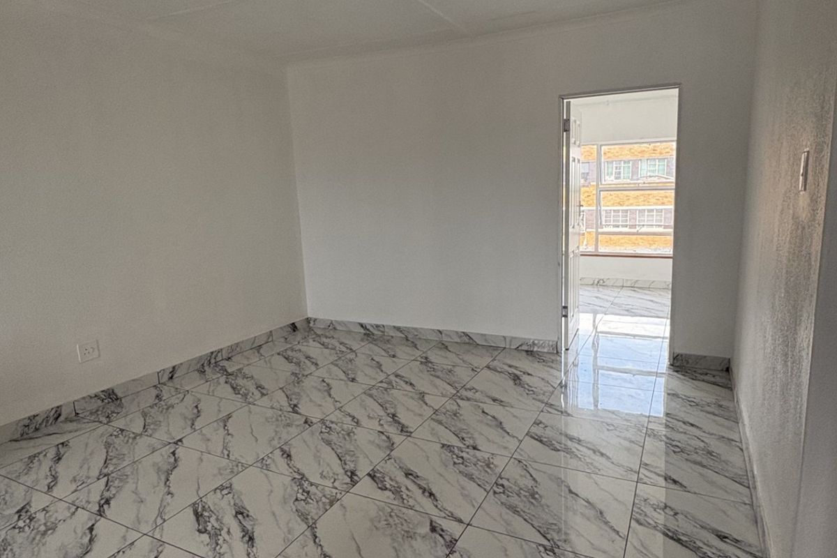 A bright, empty bedroom featuring modern marble-effect floor tiles and a doorway leading to an adjacent balcony area.