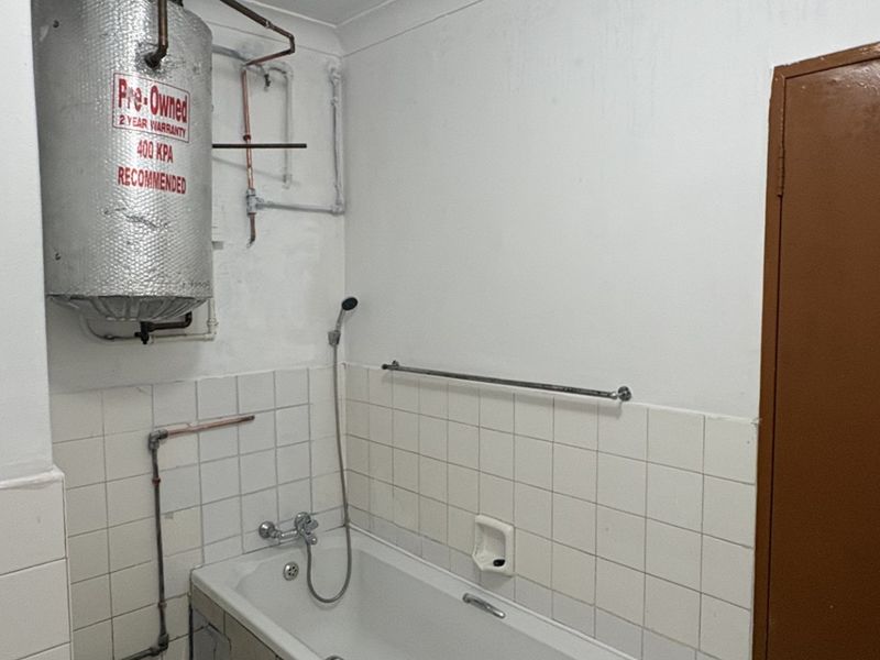 A functional bathroom featuring a bathtub with shower mixer, tiled splashback, and an exposed electric water heater unit.