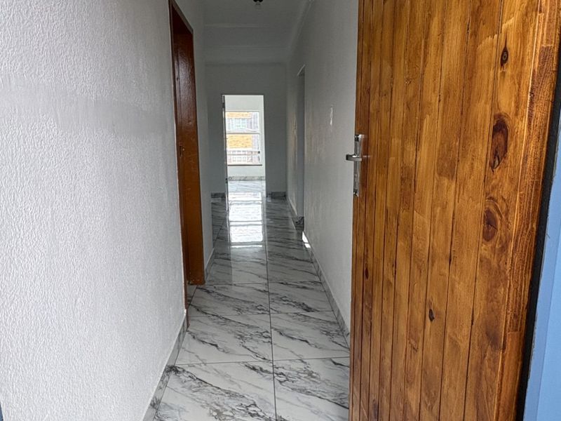 A bright, empty hallway featuring modern marble-look floor tiles and a natural wood door, leading towards an open doorway at the end.