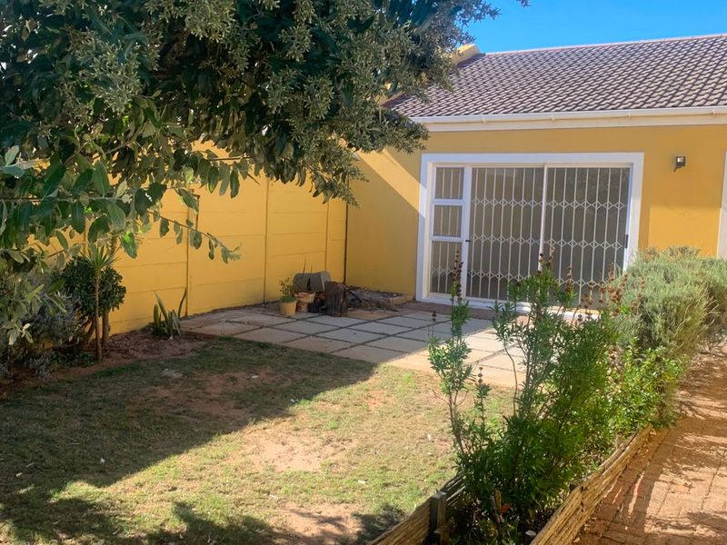 A rear exterior view featuring a paved patio area, sliding glass doors with security gates, and a small garden space with a boundary wall.