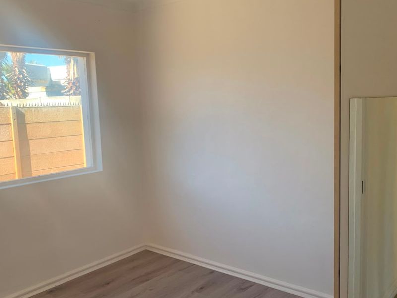 A bright, empty bedroom featuring modern laminate flooring, a built-in cupboard, and a window overlooking the exterior area.