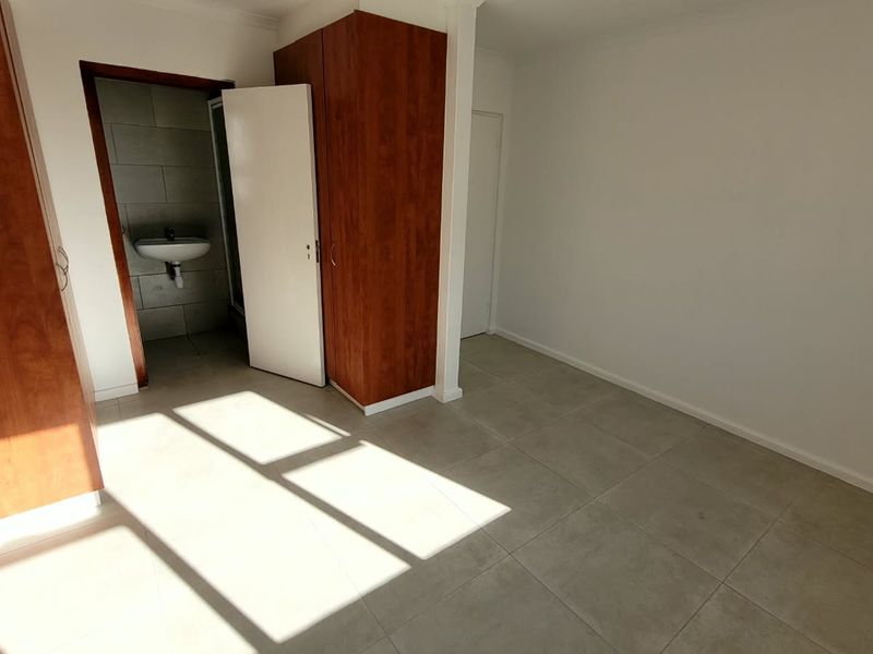 A bright, empty bedroom featuring neutral tiled flooring, built-in wooden cupboards, and direct access to an en-suite bathroom.