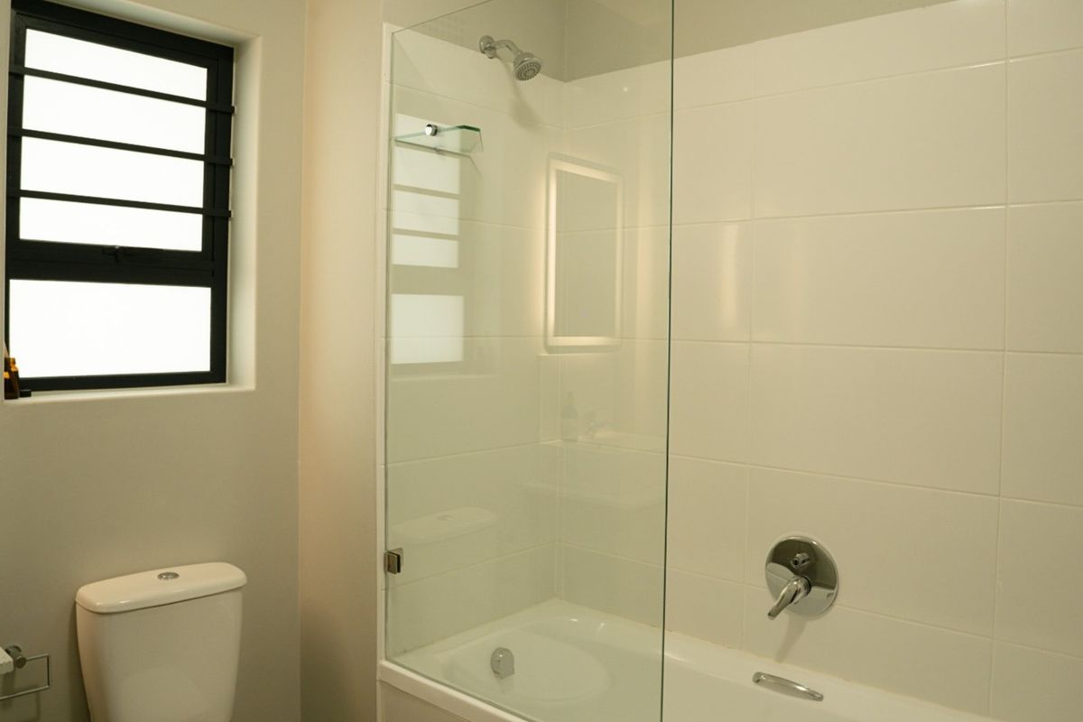 A clean, modern bathroom featuring a white bathtub with a glass shower screen, a wall-mounted toilet, and a vanity basin.