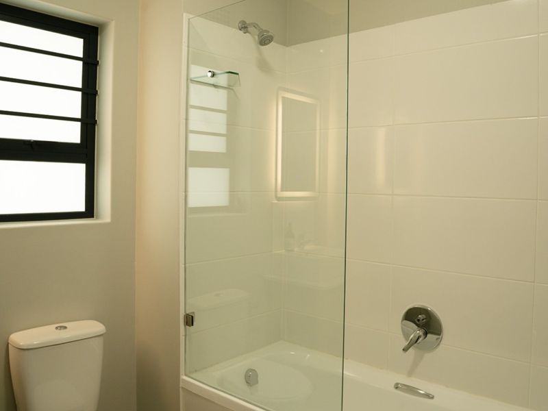 A clean, modern bathroom featuring a white bathtub with a glass shower screen, a wall-mounted toilet, and a vanity basin.