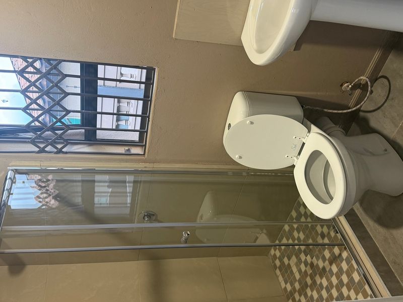 A functional bathroom featuring a shower enclosure, pedestal basin, and toilet, with tiled flooring and a security-gated window.