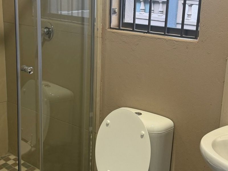A functional bathroom featuring a shower enclosure, toilet, and washbasin, with a window fitted with security bars.