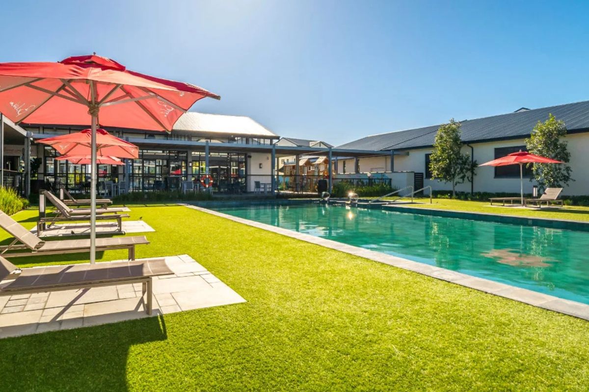 A well-maintained outdoor swimming pool area featuring a paved surround, artificial lawn, and a covered patio space for relaxation.