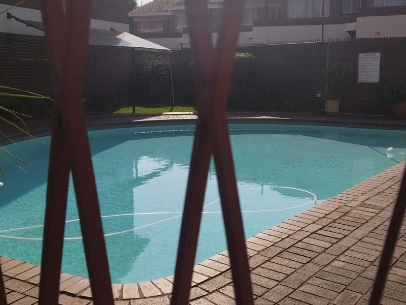 A communal swimming pool area featuring brick paving and a surrounding boundary wall, viewed through a metal security fence.