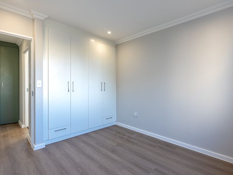 A bright, empty bedroom featuring modern laminate flooring, built-in white cabinetry, and crown molding, with an adjacent hallway showing additional storage.