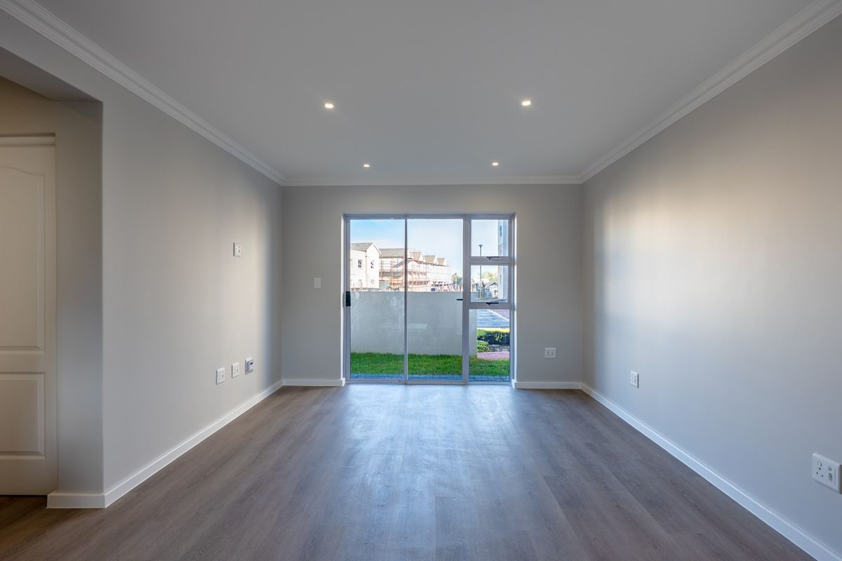 A bright, empty living room featuring modern laminate flooring, recessed downlights, crown molding, and a sliding glass door leading to an outdoor area.