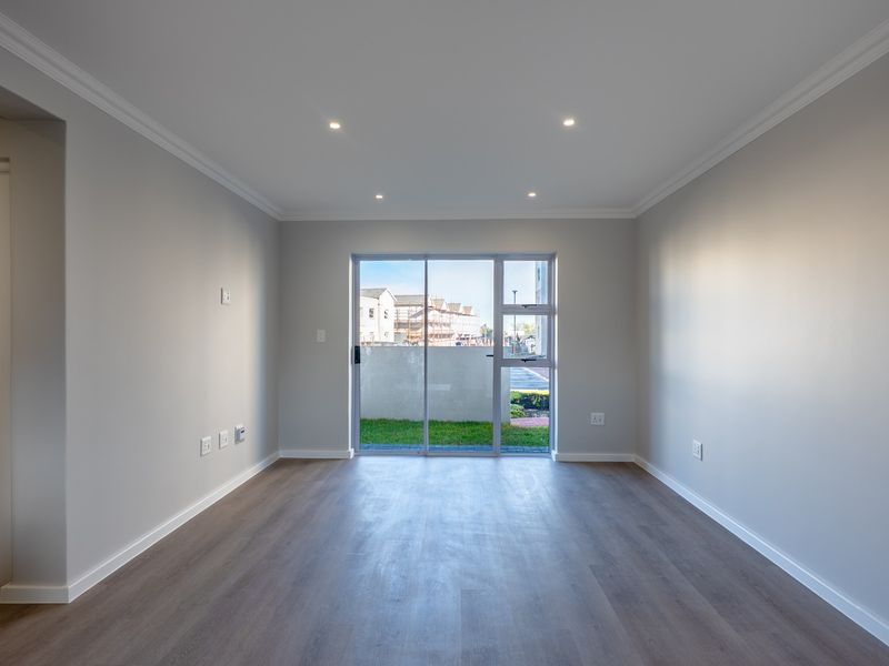 A bright, empty living room featuring modern laminate flooring, recessed downlights, crown molding, and a sliding glass door leading to an outdoor area.