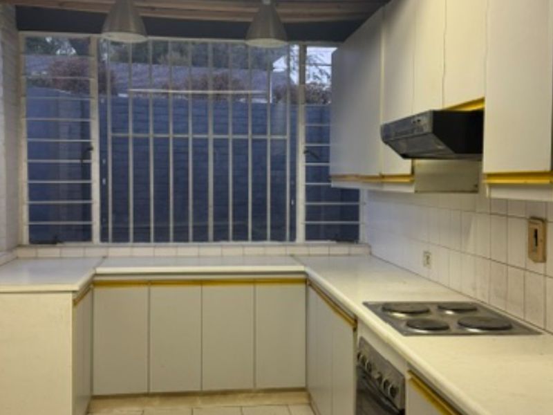 A dated kitchen featuring white cabinetry, an electric hob, built-in oven, and extractor fan, with security bars on the windows.