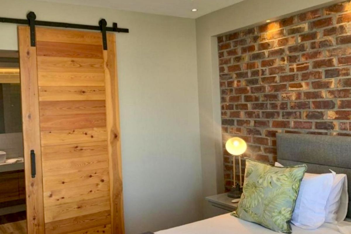 A modern bedroom featuring an exposed brick accent wall, recessed lighting, and a rustic timber sliding barn door leading to an en-suite bathroom.