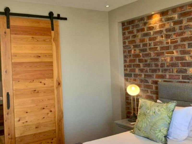 A modern bedroom featuring an exposed brick accent wall, recessed lighting, and a rustic timber sliding barn door leading to an en-suite bathroom.