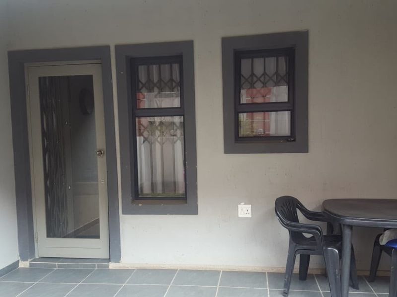 A covered patio area featuring tiled flooring, dark-framed windows with security bars, and a glass-panelled entrance door.