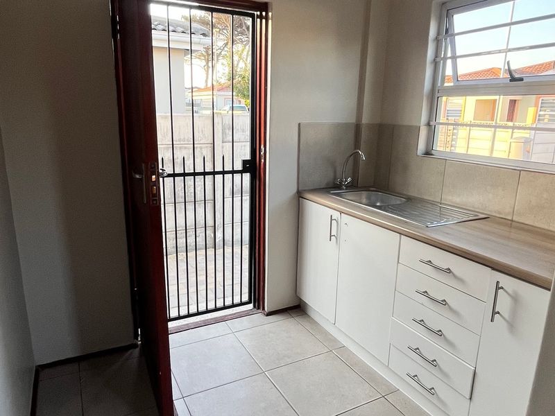 A clean and functional laundry area featuring built-in white cabinetry, a stainless steel sink, tiled splashbacks, and direct access to the exterior via a security gate.