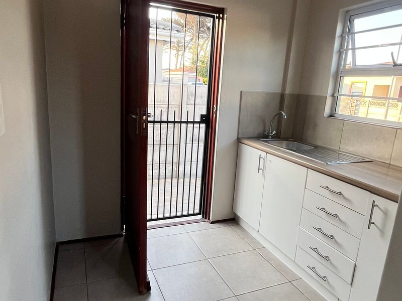 A clean and functional scullery area featuring white built-in cabinetry, a stainless steel sink, tiled splashback, and direct access to the exterior via a security gate.