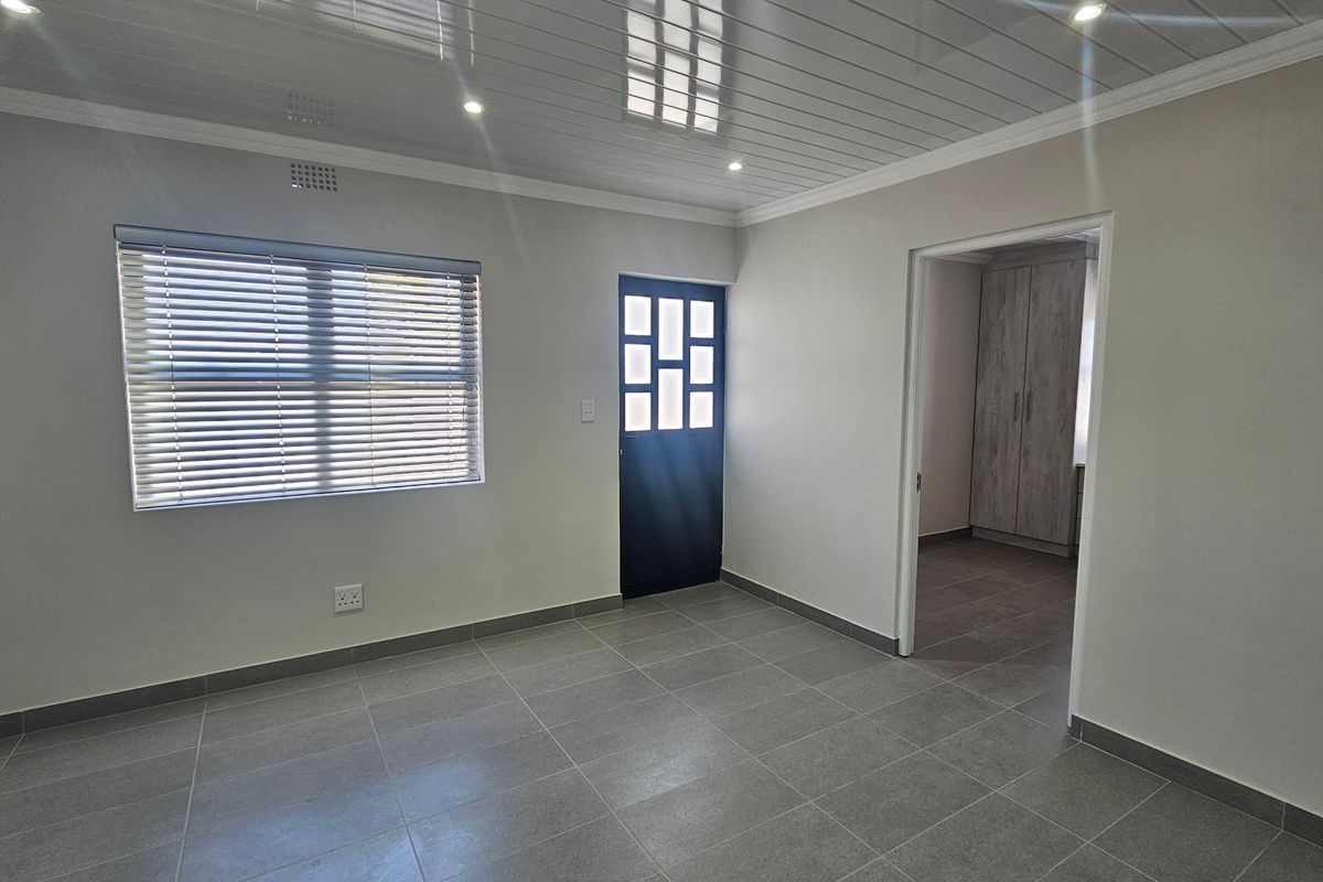 A bright, empty living area featuring modern tiled flooring, a clean PVC ceiling with recessed lighting, and a view into an adjacent room with built-in cupboards.