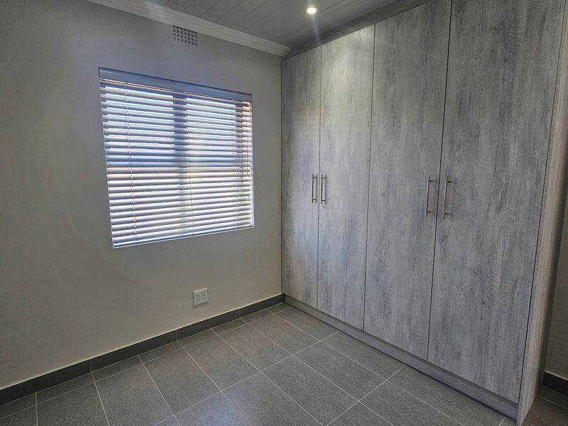 A clean, modern bedroom featuring neutral tiled flooring, built-in wood-grain cupboards, and a window fitted with venetian blinds.