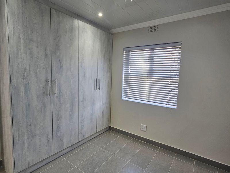 A clean, modern bedroom featuring neutral tiled flooring, built-in wood-grain cupboards, and venetian blinds, offering a bright and functional living space.