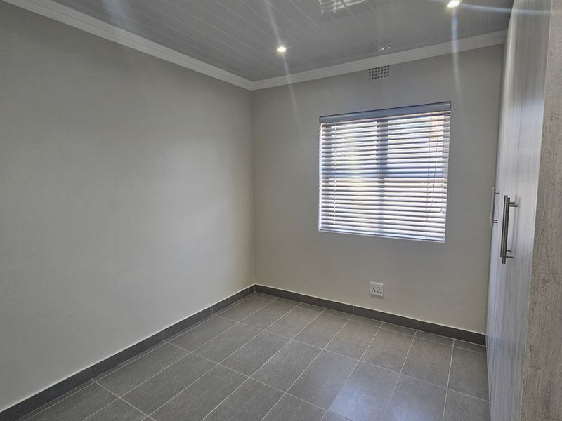 A clean, empty bedroom featuring modern grey floor tiles, built-in cupboards, recessed ceiling lighting, and venetian blinds.