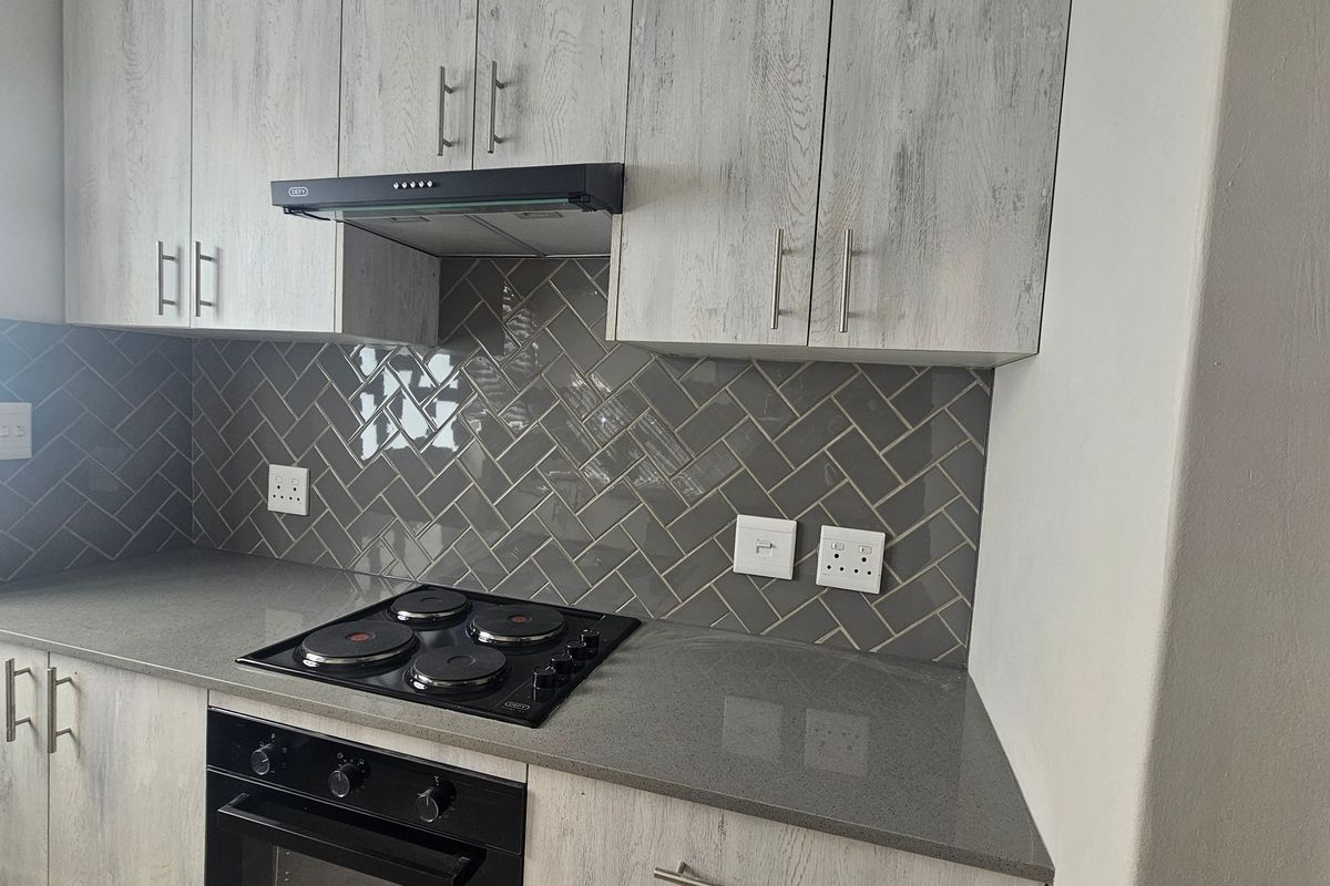 Modern kitchen featuring light wood-grain cabinetry, stone countertops, a sleek electric hob with extractor fan, and a stylish grey herringbone tile backsplash.