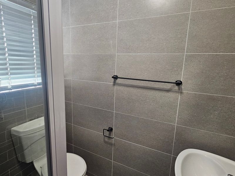 A modern, compact bathroom featuring neutral grey wall tiles, a wall-mounted basin, and contemporary black hardware fixtures.