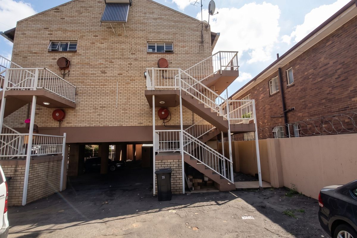 Rear view of a multi-story brick apartment building featuring external staircases, metal railings, fire safety equipment, and a covered ground-level parking area.