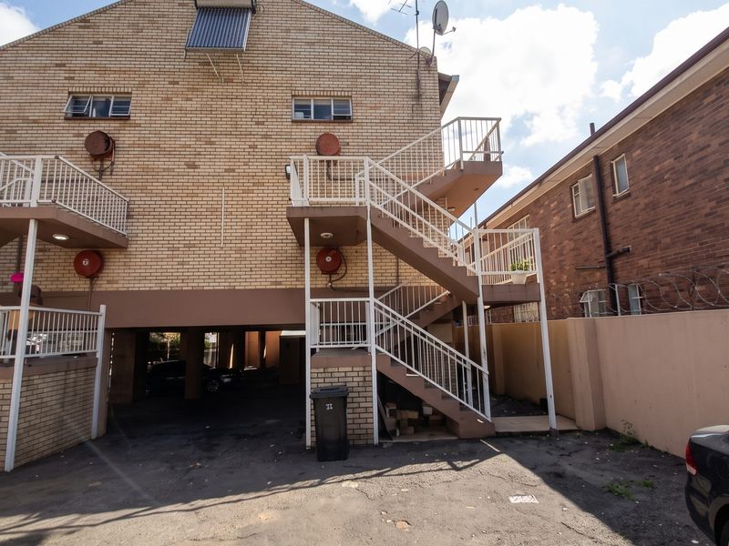 Rear view of a multi-story brick apartment building featuring external staircases, metal railings, fire safety equipment, and a covered ground-level parking area.