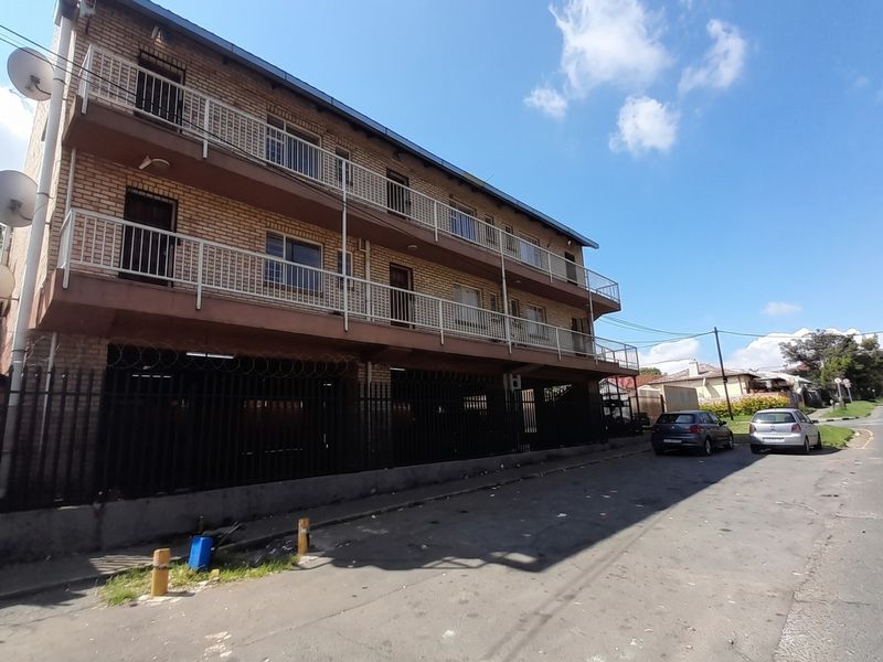 Exterior view of a multi-story brick apartment building featuring balconies with metal railings and perimeter security fencing.
