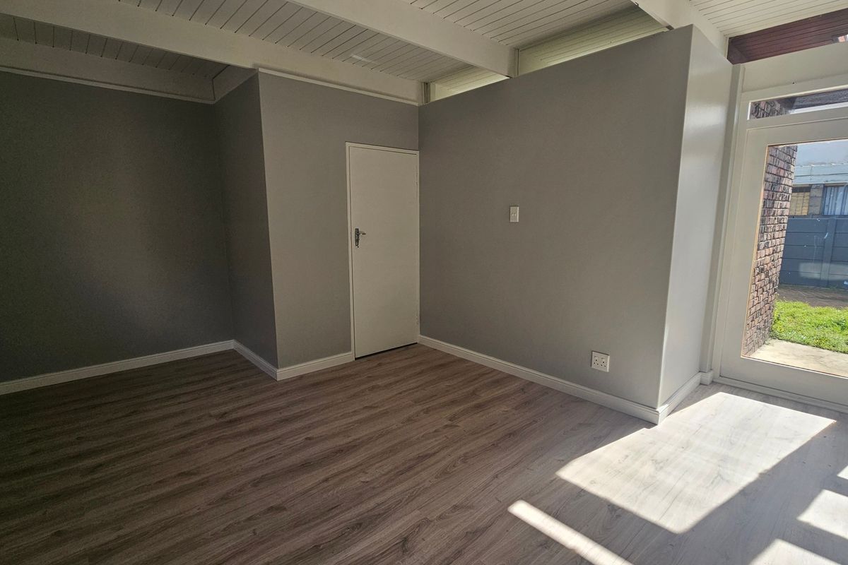 A bright, empty living area featuring modern laminate flooring, a white exposed beam ceiling, and a glass-paneled door leading to the exterior.