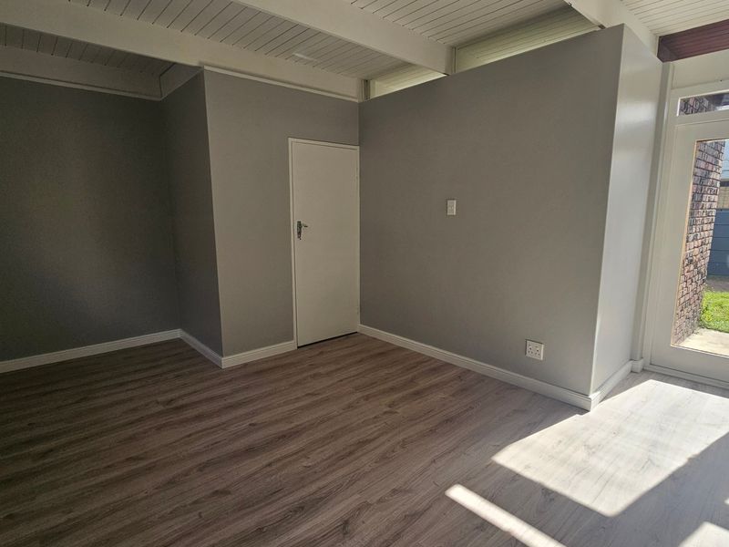 A bright, empty living area featuring modern laminate flooring, a white exposed beam ceiling, and a glass-paneled door leading to the exterior.