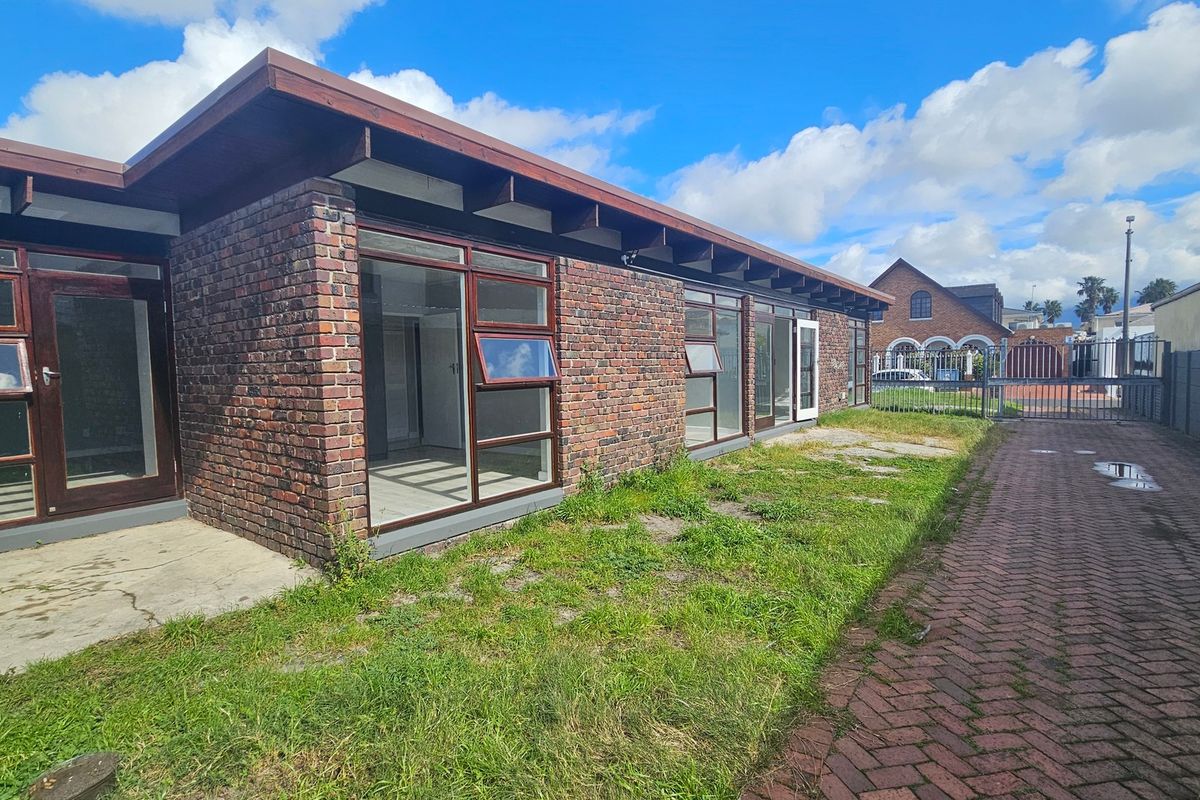 Rear view of a brick house featuring large glass doors, a timber roof structure, and a paved driveway leading to a metal security gate.