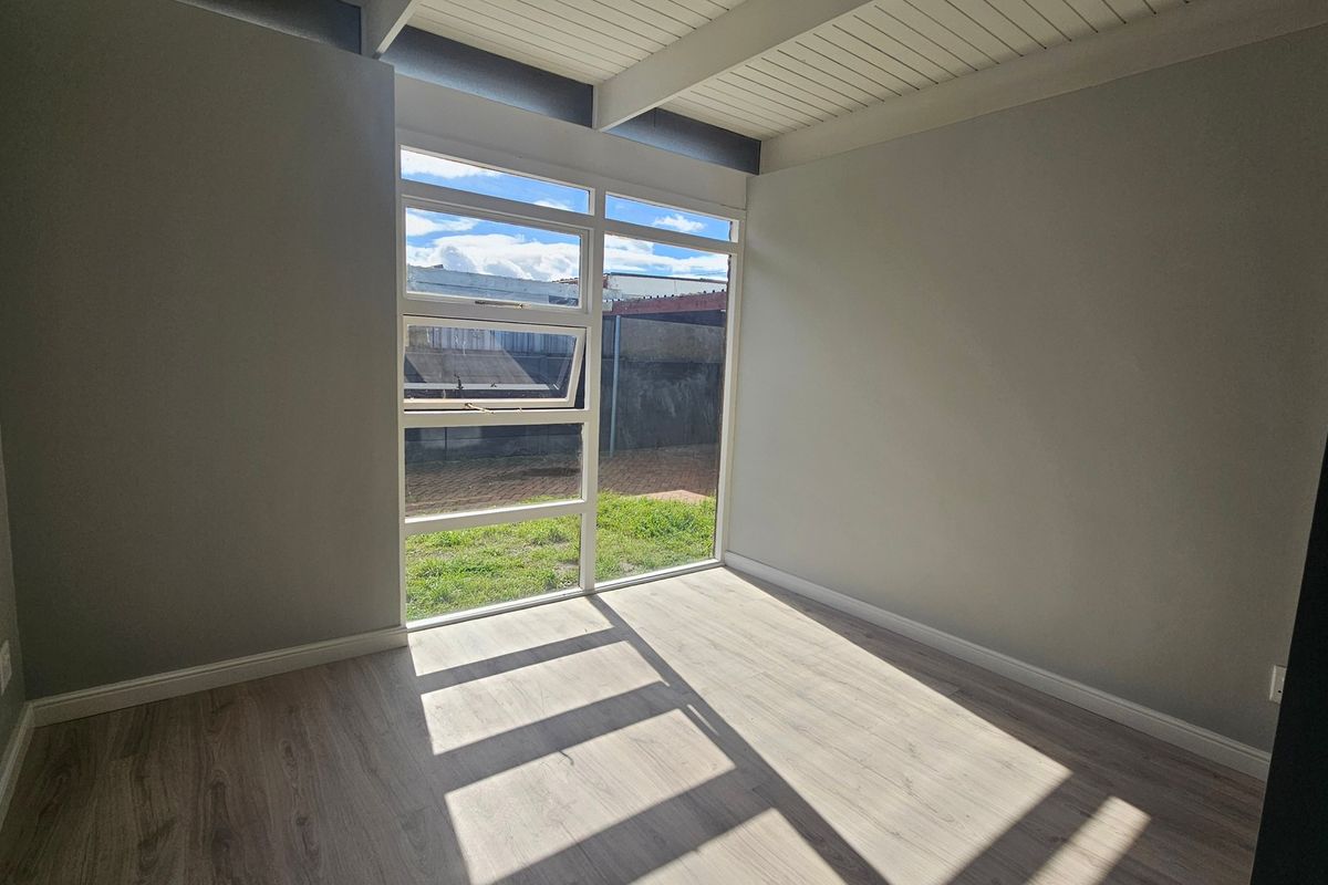 A bright, empty bedroom featuring modern laminate flooring, exposed beam ceilings, and a large window providing natural light and garden views.