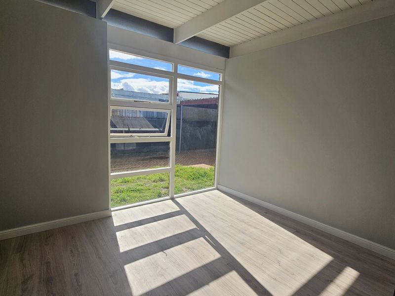 A bright, empty bedroom featuring modern laminate flooring, exposed beam ceilings, and a large window providing natural light and garden views.
