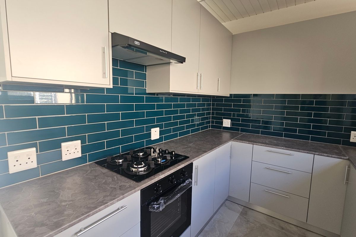 A modern, clean kitchen featuring light grey cabinetry, dark stone-look countertops, a gas hob, built-in oven, and a striking teal subway tile backsplash.