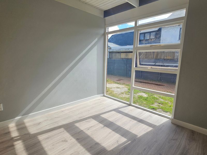 A bright, empty bedroom featuring modern laminate flooring, neutral walls, and a large window providing natural light and garden views.