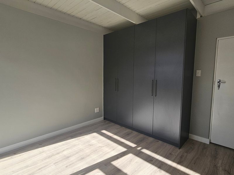 A bright, empty bedroom featuring modern dark grey built-in cupboards, light wood-look laminate flooring, and an exposed beam ceiling.