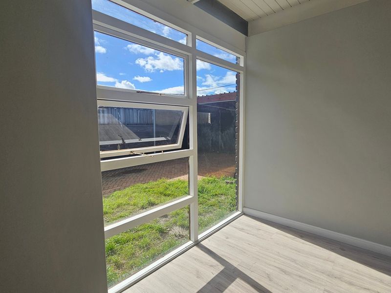 A bright, empty bedroom featuring modern laminate flooring, clean neutral walls, and a large window providing natural light and garden views.