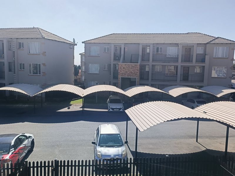 Exterior view of a residential flat complex featuring covered parking bays, asphalt driveway, and secure perimeter fencing.