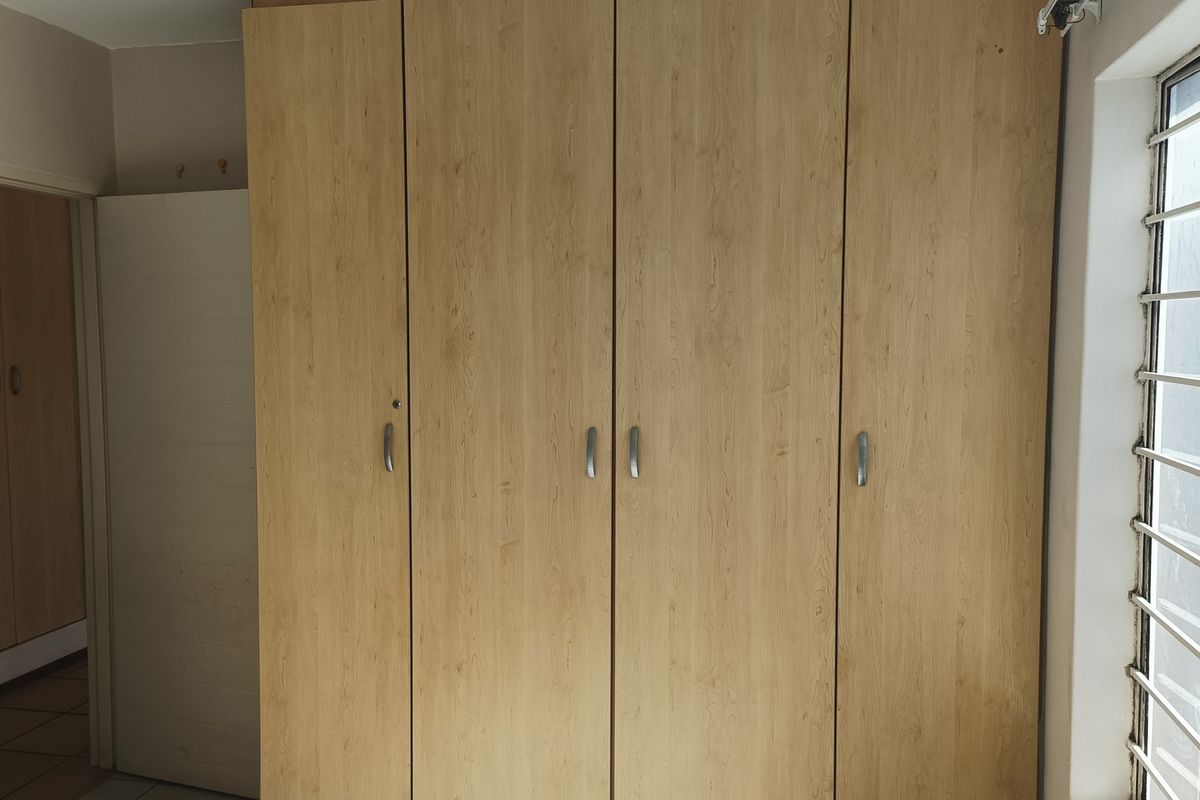 A bedroom featuring light wood-finish built-in cupboards and a window fitted with security bars.