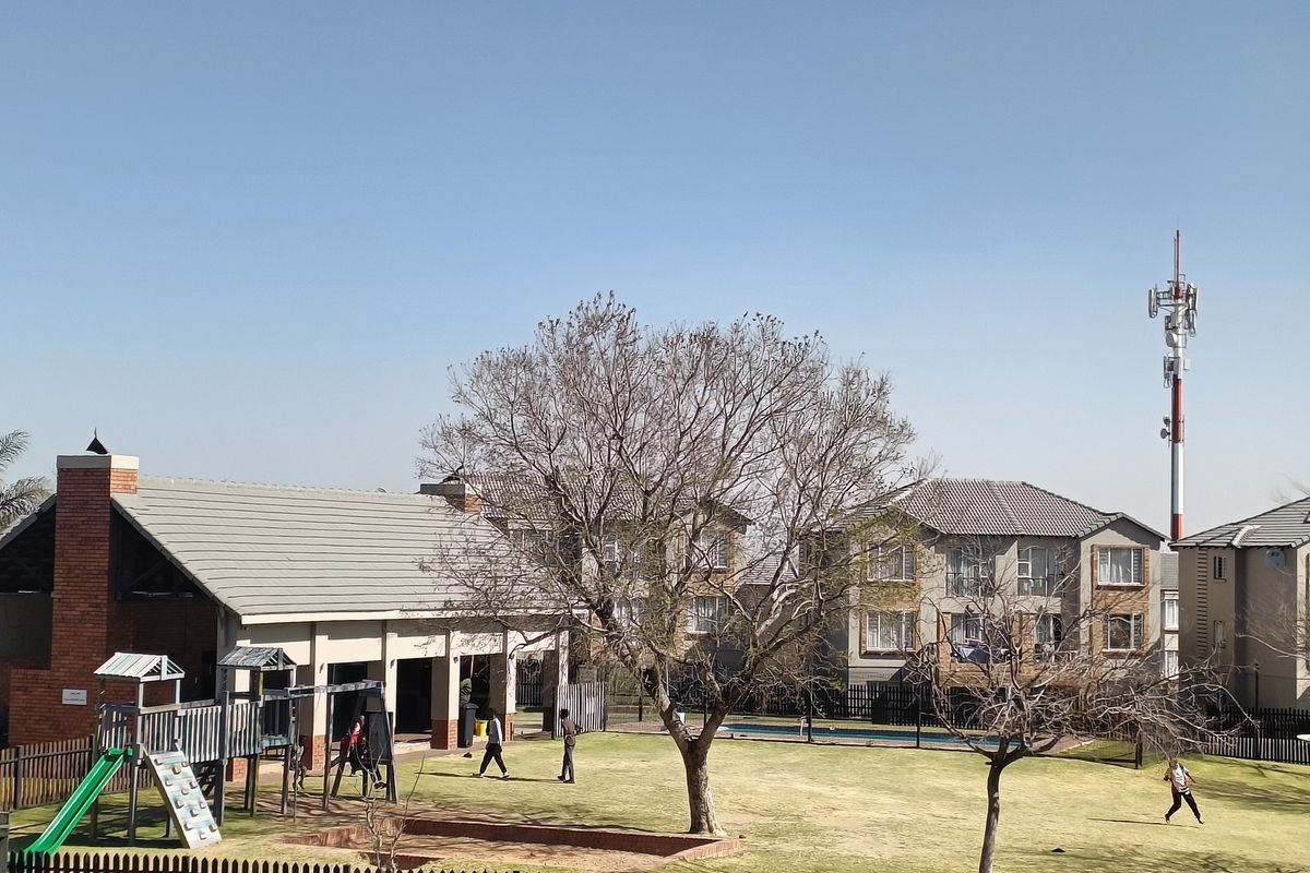 View of a communal garden area featuring a brick-built clubhouse structure, playground equipment, and well-maintained lawn within a residential complex.