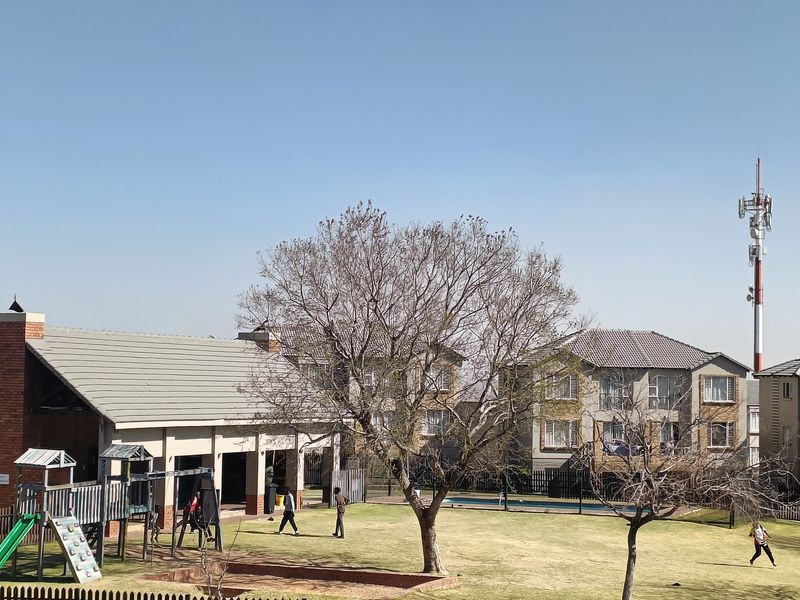 View of a communal garden area featuring a brick-built clubhouse structure, playground equipment, and well-maintained lawn within a residential complex.