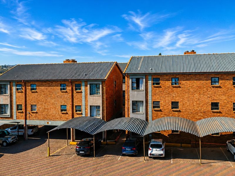 Aerial view of a multi-story brick apartment complex featuring covered parking bays and a paved driveway area.
