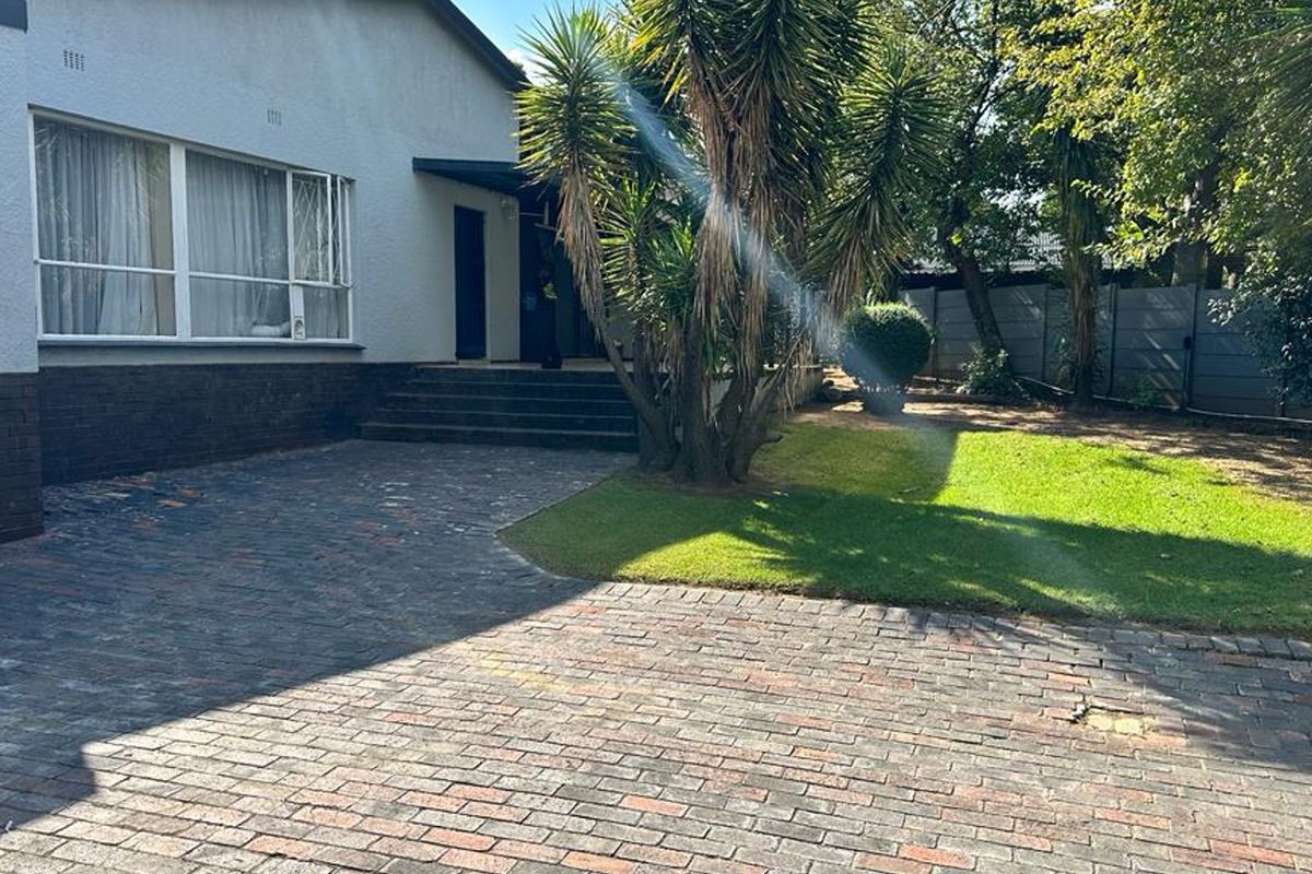 Rear exterior view featuring a brick paved driveway, concrete entrance steps, and a landscaped garden area with mature trees.