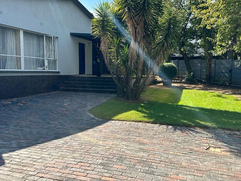 A rear exterior view of the cottage featuring a brick-paved driveway, concrete entrance steps, a well-maintained lawn, and mature garden trees.