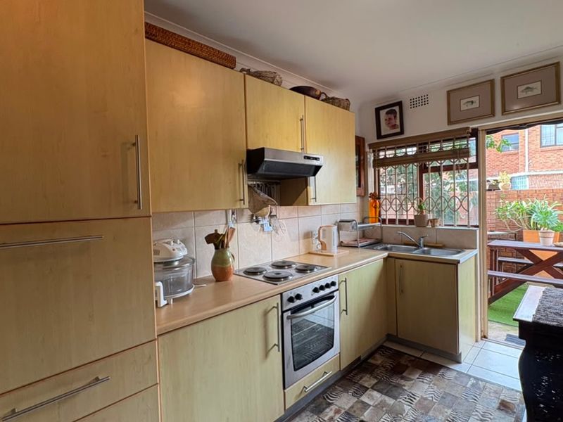 Functional kitchen featuring light wood-finish cabinetry, integrated electric hob and oven, tiled backsplash, and direct access to an outdoor patio area.