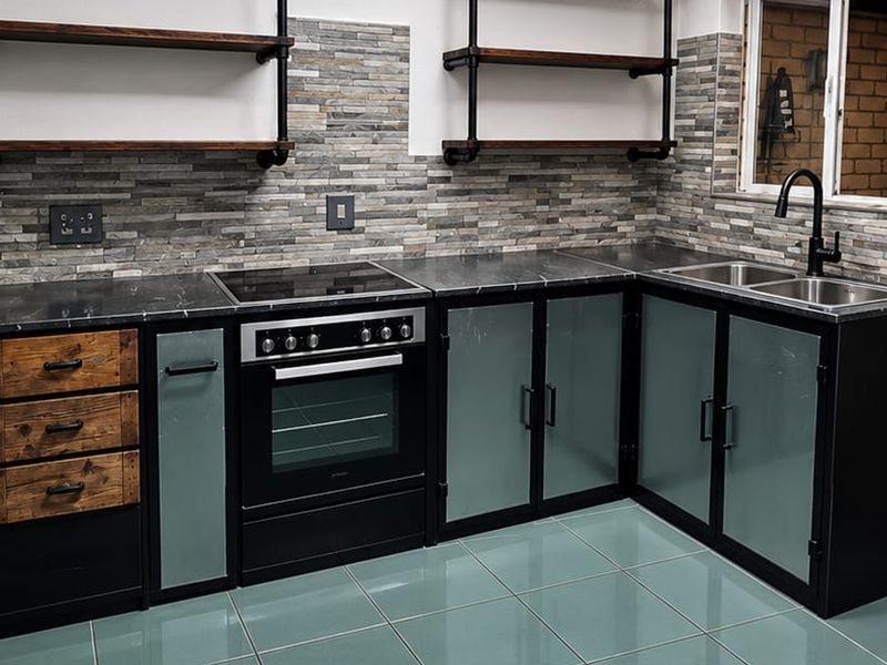 Modern industrial-style kitchen featuring dark cabinetry, stone-cladding backsplash, built-in oven with hob, and stainless steel sink with black fixtures.